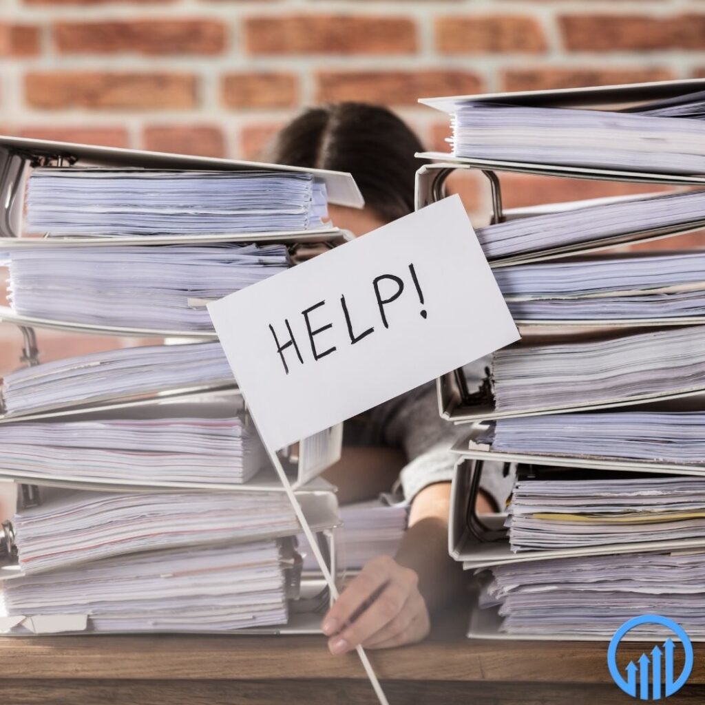 office worker stuck behind two piles of binders full of work, holding up a sign that says "help!"